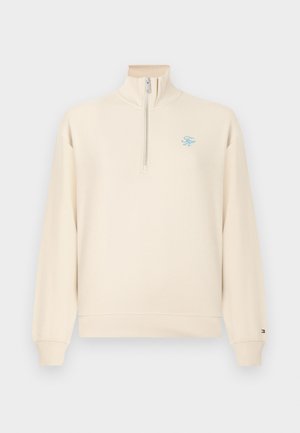 Pull à manches longues couleur crème avec une fermeture éclair sur un quart, poignets et ourlet côtelés, et petit logo brodé bleu sur le côté gauche de la poitrine.