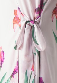 Le tissu de cette robe florale rose clair arbore des fleurs violettes et roses vives accompagnées de feuilles vertes, mettant en avant un détail de taille nouée et une texture douce et fluide.