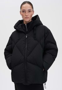 Doudoune noire avec capuche, mettant en avant un design matelassé et une fermeture éclair à l'avant. Dispose de bretelles ajustables et d'une coupe ample pour le confort.