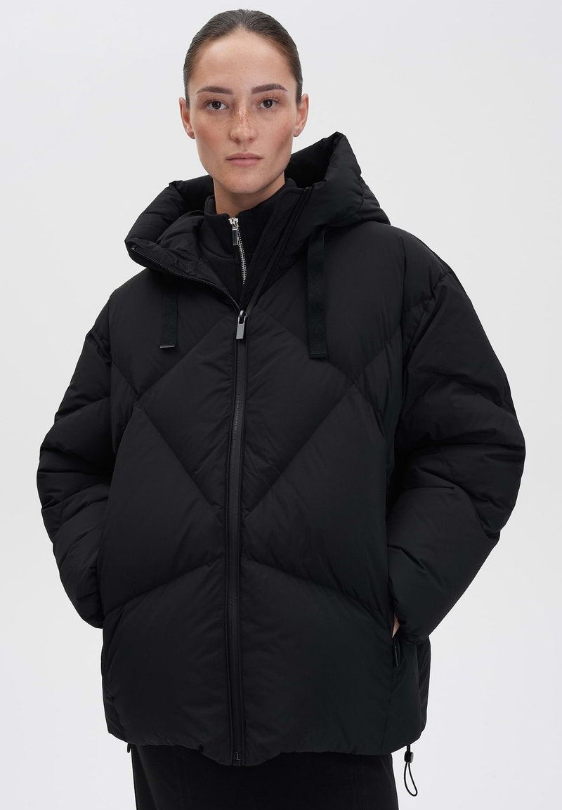 Doudoune noire avec capuche, mettant en avant un design matelassé et une fermeture éclair à l'avant. Dispose de bretelles ajustables et d'une coupe ample pour le confort.