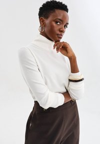 Kobieta z krótkimi kręconymi włosami, w złotych kolczykach w kształcie kół, białym swetrze z wysokim kołnierzem, ciemnych spodniach o wysokim stanie i bransoletkami na nadgarstku.