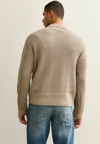 Mężczyzna w beżowym prążkowanym swetrze dzianinowym i niebieskich dżinsach, stojący tyłem do kamery na jednolitym tle.