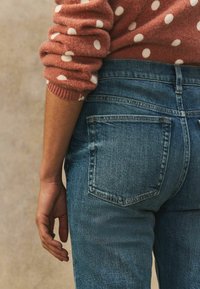 Haalistuneet siniset farkut, joissa on keskipitkä vyötärö, kaksi takataskua ja ommeltuja yksityiskohtia. Kuvioitu, pisteitä sisältävä neulepaita kurkistaa esiin.