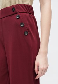 Bordeaux rode hoge taille broek met een gladde textuur, voorzien van een brede tailleband en vier zwarte knopen aan de zijkant.