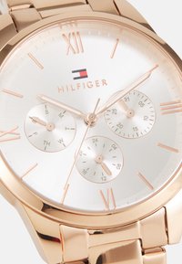 Reloj metálico color oro rosado con una esfera plateada, que presenta tres subesferas, números romanos y el logo "HILFIGER" en el centro.