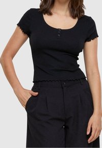 Černý žeberkový crop top s krátkými rukávy, zvlněnými okraji a kulatým výstřihem. Obsahuje tři ozdobné knoflíky vpředu.