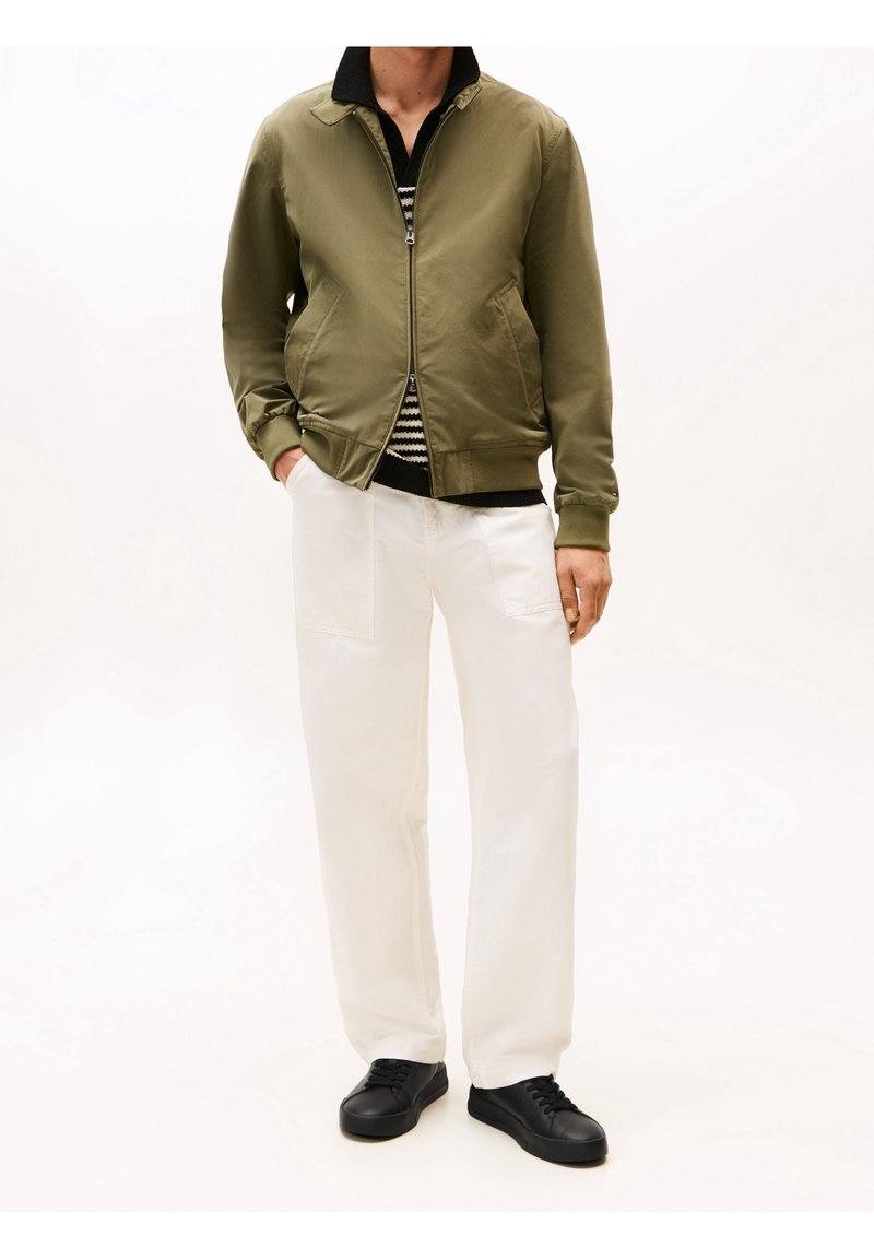 Homme portant une veste zippée vert olive, un pull rayé noir et blanc, un pantalon blanc et des baskets noires, debout avec une main dans la poche.