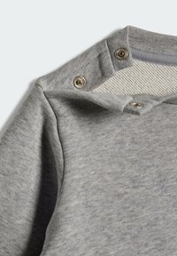 Felpa grigia con colletto a coste, tessuto strutturato, cuciture visibili e bottoni a pressione argentati sulla spalla per una facile vestibilità.