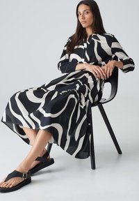 Schwarzes und weißes gemustertes Hemdkleid mit einem gestuften Saum, gefertigt aus einem leichten Stoff. Getragen mit schwarzen flachen Sandalen, sitzend auf einem schwarzen Stuhl.