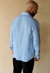 Chemise à manches longues rayée bleu clair avec un col classique, une texture douce, un ourlet droit et des poignets boutonnés, portée avec un pantalon noir.