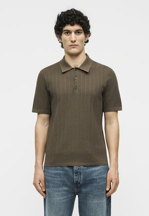 Ένας νεαρός άνδρας στέκεται απέναντι φοράει καφέ κοντομάνικη πλεκτή πόλο μπλούζα με κουμπιά και μπλε τζιν μπροστά σε απλό φόντο.