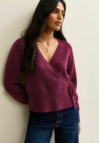 Maglione avvolgente borgogna con maniche lunghe a sbuffo, tessuto a maglia testurizzato e dettagli con nodo laterale, abbinato a jeans in denim blu scuro.