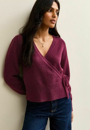 Pull enveloppant bordeaux avec de longues manches bouffantes, en tissu tricoté texturé, et un détail de lien sur le côté, associé à un jean en denim bleu foncé.