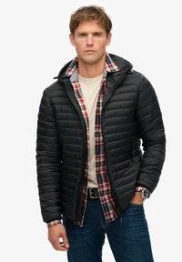 Sort puffet jakke med hætte, der har et vandret quiltet mønster. Båret over en rød og blåternet flannelskjorte, parret med jeans.