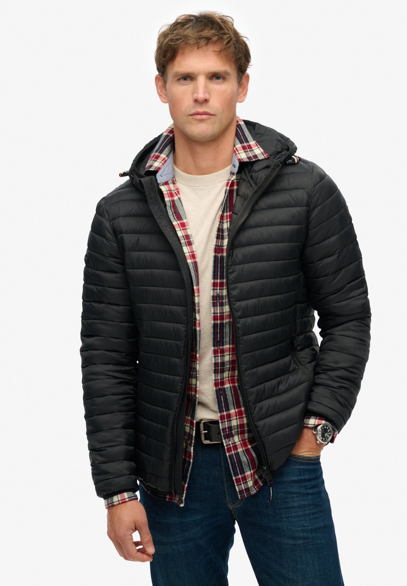 Sort puffet jakke med hætte, der har et vandret quiltet mønster. Båret over en rød og blåternet flannelskjorte, parret med jeans.