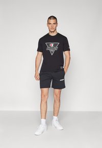 Svart t-shirt med kort ärm och en triangellogo samt texten "CHAMPION", tillsammans med svarta shorts och vita sneakers, på en enfärgad bakgrund.