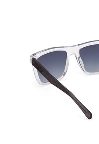 Gafas de sol transparentes con lentes gris degradado, diseño angular y elegantes brazos negros con el nombre de la marca "GUESS" embellecido.