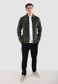 Reißverschluss-Front, grüne Cordjacke mit kragenförmigem Ausschnitt, kombiniert mit einem weißen T-Shirt und schwarzen Hosen, getragen mit hellbeigen Sneakern.