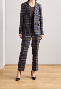 Navy ja ruskea ruutukuosi puku, joka on valmistettu rakenteellisesta kankaasta. Mukana on räätälöity bleiseri, jossa on taskut, ja istuvat housut. Mustat korkokengät.