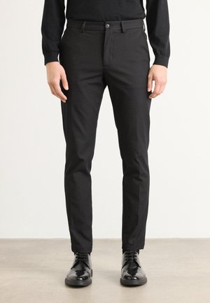 Black, slim-fit trousers with a smooth texture, featuring a zipper fly and button, paired with polished black shoes.