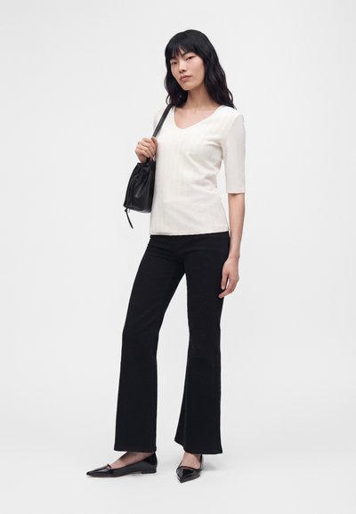 Femme debout portant un haut blanc texturé, un pantalon noir évasé, des ballerines noires pointues et un sac à épaule noir sur un fond uni.