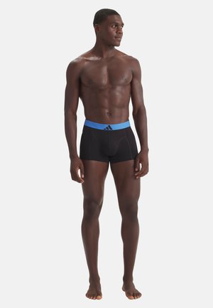 Mandlig model stående barfodet iført sorte boxershorts med blå linning og Adidas-logo, der ser let mod højre på en hvid baggrund.