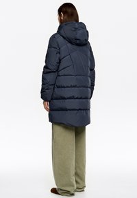 Granatowa pikowana kurtka puchowa z kapturem, pikowanym wzorem i wydłużonym krojem. Modelka ma na sobie luźne oliwkowozielone spodnie i brązowe obuwie.