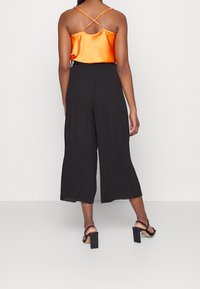 Osoba ubrana w pomarańczowy satynowy top na ramiączkach, czarne szerokie spodnie do kostek i czarne sandały na obcasie, stojąca na tle białego tła.