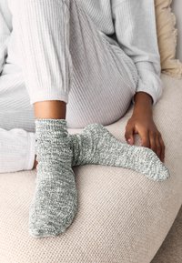 Chaussettes tricotées dans un mélange vert et blanc, présentant un design lâche. Portées avec une tenue côtelée gris clair sur une surface texturée claire.