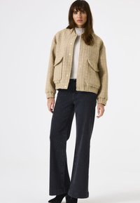 Femme portant une veste à carreaux beige avec de grandes poches, un pull à col roulé crème, un pantalon large noir avec des éclats subtils, et des bottes noires.