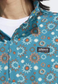Turkosblå skjorta med krage och ett solmönster i orange, vitt och ljusblått. Har en etikett från "adidas Adventure" på vänster sida.