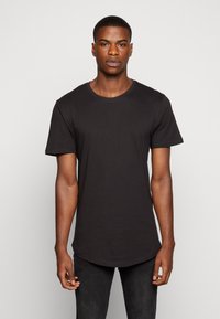 Schwarzes Baumwoll-T-Shirt mit kurzen Ärmeln, einem runden Ausschnitt und einem leicht abgerundeten Saum. Getragen mit dunklen Jeans, vor einem schlichten weißen Hintergrund.