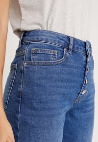 Jean en denim bleu foncé taille haute avec une fermeture à cinq boutons, un léger effet usé, et des poches avant et arrière. Couture de couleur contrastante.
