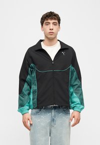 Jeune homme portant une veste zippée noire et turquoise avec un logo Puma, un sous-pull blanc et un jean bleu clair avec des logos Puma.