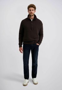 Donkerbruin getextureerd sweater met hoge kraag en kwart-rits, in combinatie met donkerblauwe jeans en witte sneakers met tan kleurige zolen.