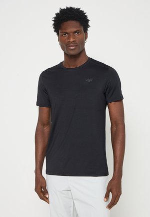 Hombre con el pelo corto y rizado, que lleva una camiseta negra ajustada de manga corta y pantalones de color claro, de pie frente a un fondo liso.