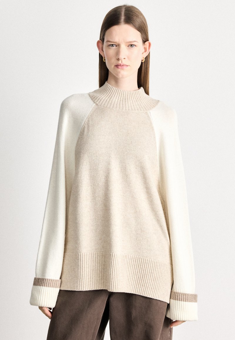 Μπεζ και εκρού πλεκτό πουλόβερ με ψηλό γιακά, ριμπ τελείωμα και φαρδιά μανίκια με καφέ λεπτομέρεια. Μαλακή υφή και άνετη γραμμή.