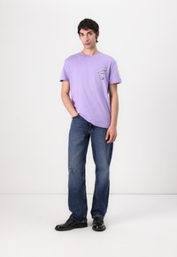 Lilla bomull t-skjorte med korte ermer og et lite grafisk trykk på venstre bryst, parret med blå jeans og svarte sko.