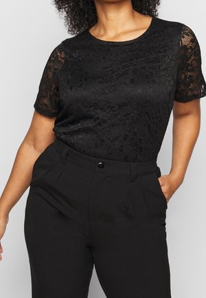 Vrouw in een zwart kanten top met korte mouwen, gestoken in hoge zwarte broek, met één hand in haar zak tegen een lichte achtergrond.