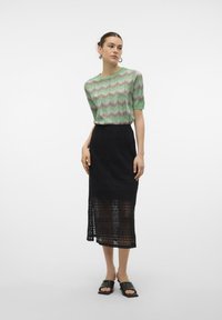 Top tricotat cu model zigzag verde și roz, cu mâneci scurte, asortat cu o fustă midi din dantelă neagră și sandale negre de tip slide.