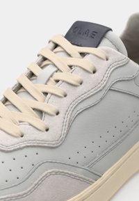 Adidași gri deschis din piele și suede, cu șireturi bej. Prezintă un design texturat, perforații subtile și un patch logo în nuanță de bleumarin.
