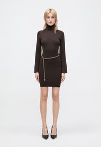 Brun ribbet strikket kjole med høj hals, lange ærmer og et guld kædebælte som accent. Kombineret med sorte spidse hæle.