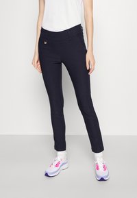 Pantaloni elasticizzati blu navy con una texture liscia, gamba affusolata e un accento metallico in vita, abbinati a scarpe da ginnastica bianche.