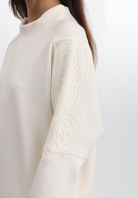 Blusa crema con colletto alto e delicati dettagli in pizzo sul maniche sinistra; texture morbida e vestibilità rilassata, senza chiusure visibili.