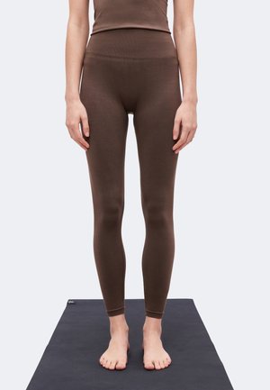 Mujer de pie descalza sobre una esterilla de yoga negra, con leggings ajustados marrones y una camiseta a juego, mostrada desde los hombros hasta los pies.