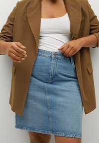 Blazer marron sur un top blanc sans manches et une jupe en denim bleue. La jupe présente une couture frontale et une texture en denim typique. Le blazer a des poches.