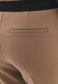 Beige broek met een gladde textuur, voorzien van een achterzak en een zwarte tailleband. De stof lijkt licht elastisch en op maat gemaakt.