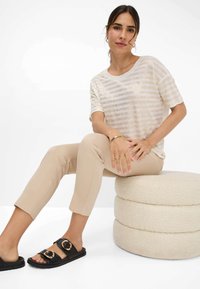 Lichtgewicht, gestreepte, crèmekleurige top met metalen accenten, gecombineerd met beige, aansluitende broek en zwarte sandalen met gouden hardware, zittend op een gestructureerde kruk.