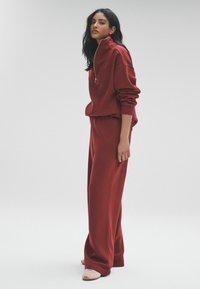 Červená mikina na zips s vysokým golierom, skombinovaná s voľnými nohavicami. Mäkká textura, ležérny dizajn, so širokými rukávmi a manžetami.