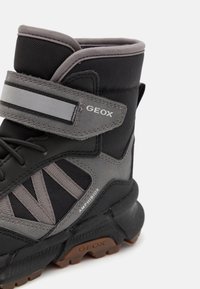 Botas impermeables en negro y gris con caña alta, que cuentan con una correa de velcro y una superficie texturizada, puntera reforzada y suela de goma.